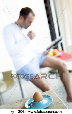 Stock Image - Man sitting on kitchen table, reading magazine, boiled egg in foreground. Fotosearch