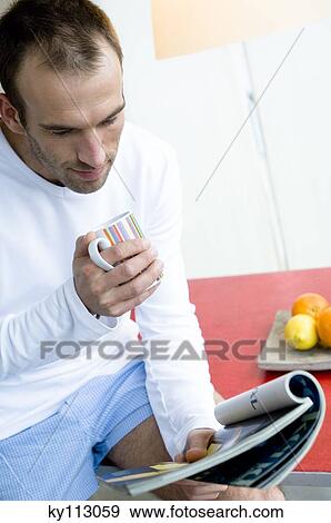 Man sitting on kitchen table, reading magazine, holding mug View Large Photo Image Stock Photo - Man sitting on kitchen table, reading magazine, holding mug. Fotosearch