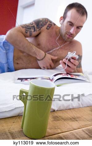 Stock Photo - Tattooed man, barechested, listening to iPod player, lying on a bed, mug in foreground. Fotosearch