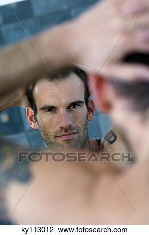 Stock Image - Tattooed man, barechested, looking in bathroom mirror. Fotosearch