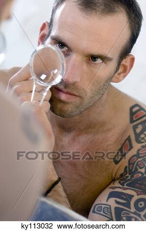 Stock Image - Tattooed man, barechested, looking in bathroom mirror, holding a magnifying glass. Fotosearch
