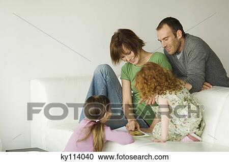 Stock Image - Couple and 2 little girls reading a magazine in a living-room. Fotosearch