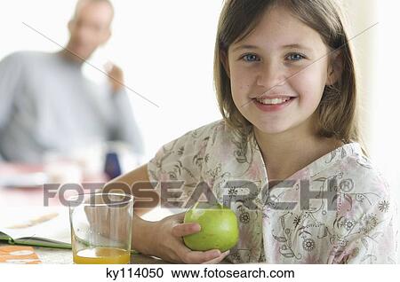 Stock Image - Portrait of a little girl eating an apple, man in the background. Fotosearch