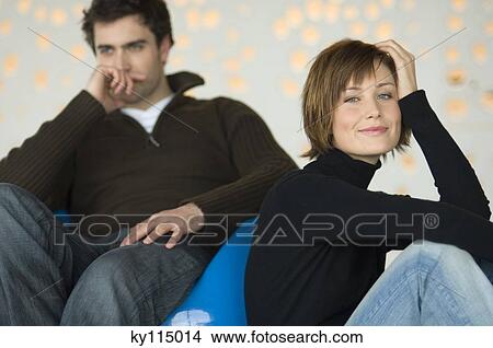 Couple sitting in living-room, woman looking at the camera, man thinking in the background View Large Photo Image Picture - Couple sitting in living-room, woman looking at the camera, man thinking in the background. Fotosearch