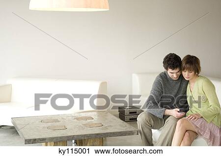 Stock Image - Couple sitting on a sofa in living-room, using mobile phone. Fotosearch