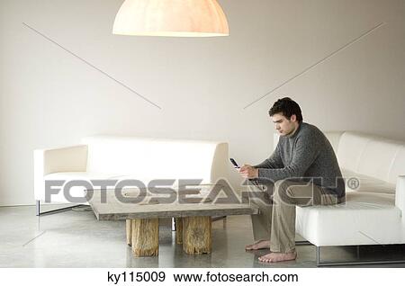 Man sitting on a sofa in living-room, using mobile phone View Large Photo Image Stock Photo - Man sitting on a sofa in living-room, using mobile phone. Fotosearch