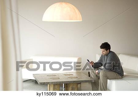 Stock Photo - Man sitting on a sofa in living-room, using mobile phone. Fotosearch