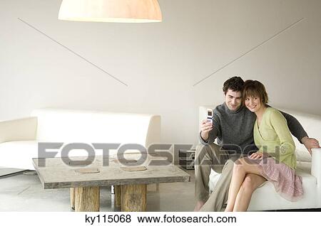 Smiling couple sitting on a sofa, taking picture with camera phone View Large Photo Image Stock Photo - Smiling couple sitting on a sofa, taking picture with camera phone. Fotosearch
