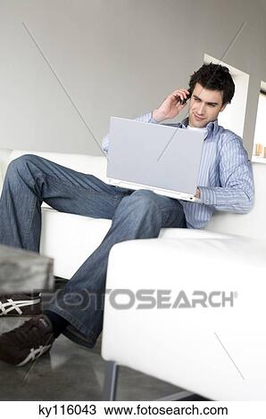 Man sitting on a sofa, using laptop computer and mobile phone View Large Photo Image Stock Image - Man sitting on a sofa, using laptop computer and mobile phone. Fotosearch