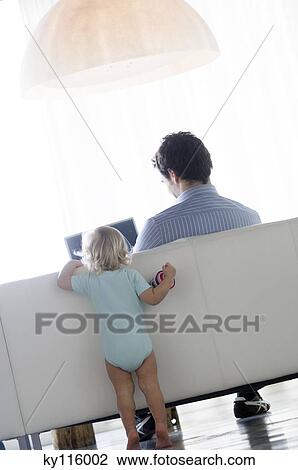 Stock Image - Rear view of little boy watching man using laptop computer. Fotosearch