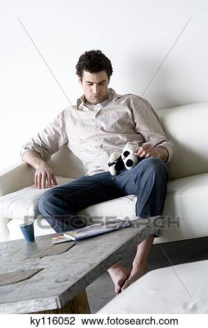 Stock Image - Sad looking man sitting on a sofa, holding stuffed dog. Fotosearch