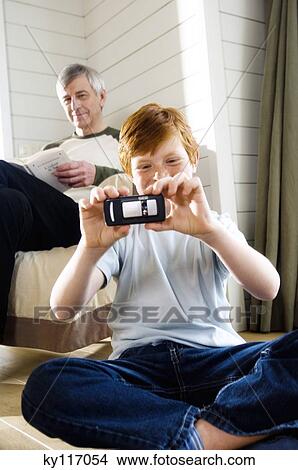 Boy sitting cross-legged, taking picture with camera phone, senior man reading in background View Large Photo Image Picture - Boy sitting cross-legged, taking picture with camera phone, senior man reading in background. Fotosearch