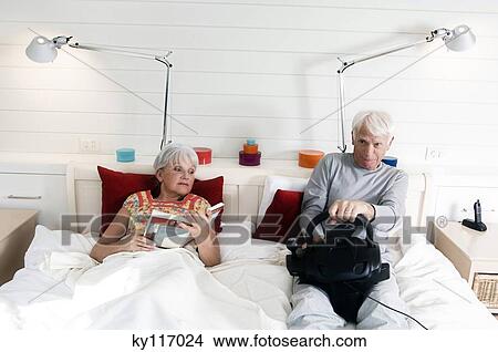 Senior couple in bed, woman reading, man playing video game View Large Photo Image Picture - Senior couple in bed, woman reading, man playing video game. Fotosearch