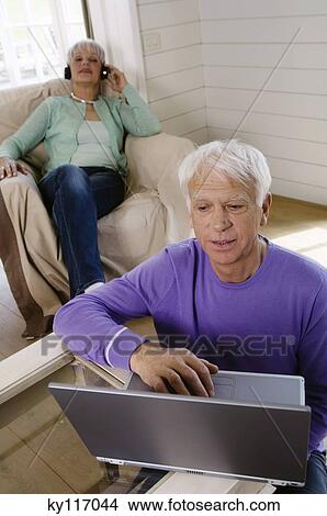 Senior couple in the living-room, man using laptop computer, woman wearing headphones in background View Large Photo Image Picture - Senior couple in the living-room, man using laptop computer, woman wearing headphones in background. Fotosearch