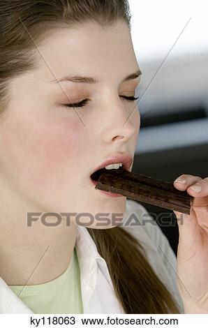 Stock Image - Portrait of a young woman eating chocolate bar, eyes shut. Fotosearch