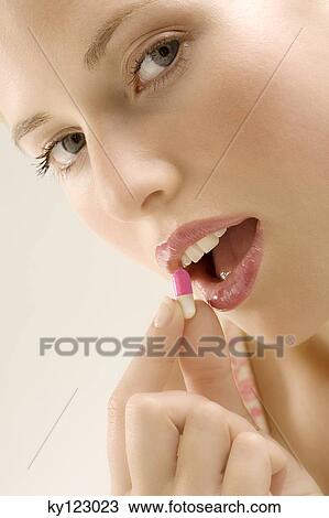 Stock Image - Portrait of a young woman opening her mouth for a capsule, indoors (studio). Fotosearch