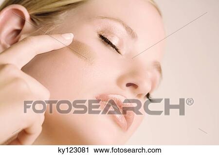 Stock Image - Woman putting make-up base on her cheek, shut eyes, indoors (studio). Fotosearch