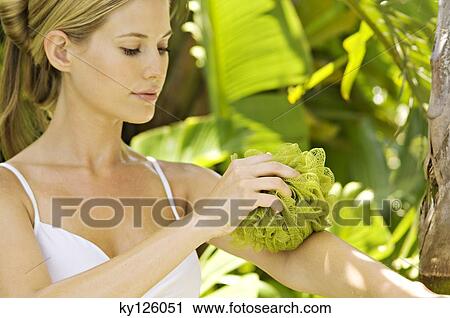 Stock Image - Portrait of a young woman cleaning her arm with a mesh sponge, outdoors. Fotosearch