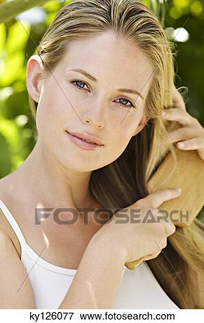 Stock Photo - Portrait of a young woman looking at the camera, brushing her hair, outdoors. Fotosearch