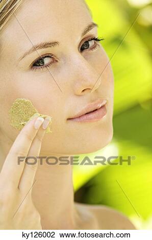 Stock Image - Young woman applying face scrub cream on her face, outdoors. Fotosearch