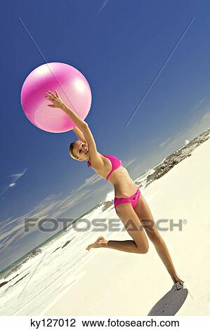 Stock Image - Young smiling woman in pink bikini on the beach, lifting a large ball. Fotosearch