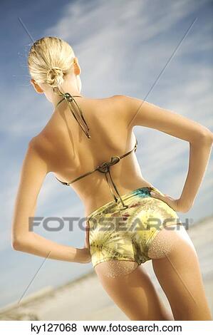 Stock Photo - Young woman in bikini standing on beach, rear view. Fotosearch