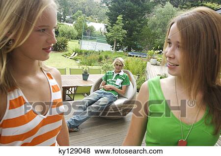 Stock Photo - 2 teenage girls looking at each other on a terrace, teenage boy sitting in background. Fotosearch