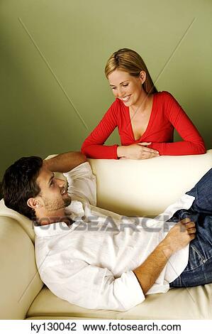 Stock Image - Young woman smiling at man lying on sofa. Fotosearch