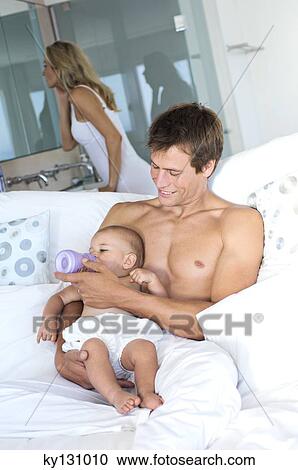 Father feeding her baby, sitting in sofa, woman in background, indoors View Large Photo Image Stock Image - Father feeding her baby, sitting in sofa, woman in background, indoors. Fotosearch