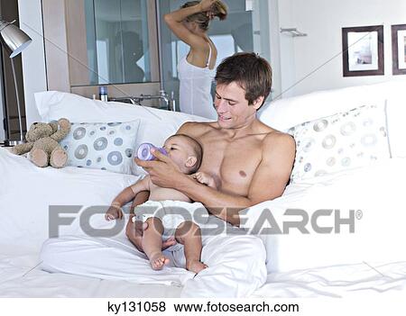 Father feeding his baby, mother in background, indoors View Large Photo Image Stock Photo - Father feeding his baby, mother in background, indoors. Fotosearch