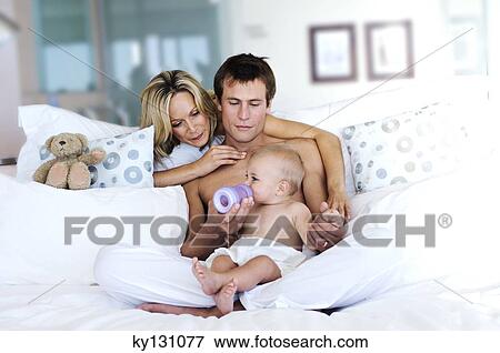 Stock Photo - Father feeding his baby, mother looking at them, indoors. Fotosearch
