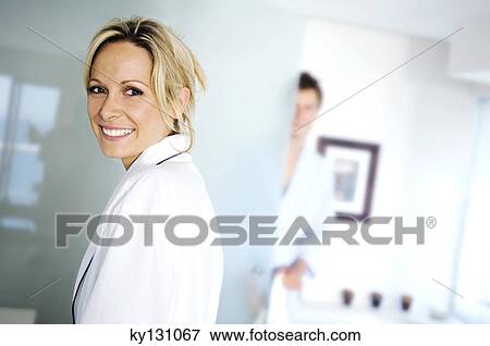 Portrait of a young woman smiling, looking at the camera, man in background, indoors View Large Photo Image Stock Photo - Portrait of a young woman smiling, looking at the camera, man in background, indoors. Fotosearch