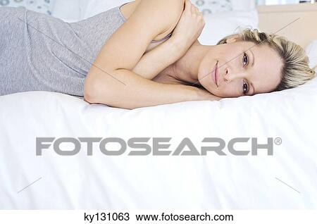 Stock Image - Young woman looking at the camera, lying on bed, indoors. Fotosearch