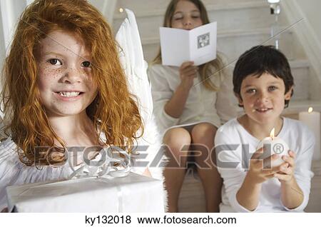 Christmas day, portrait of a little girl holding a present, sister and brother in background with card and candle, indoors View Large Photo Image Stock Photo - Christmas day, portrait of a little girl holding a present, sister and brother in background with card and candle, indoors. Fotosearch