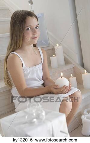 Stock Photograph - Christmas, little girl sitting indoors, holding a candle, indoors. Fotosearch