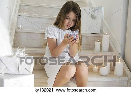 Stock Image - Little girl sitting indoors, holding a Christmas ball, indoors. Fotosearch