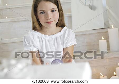 Stock Image - Little girl sitting indoors, holding a Christmas ball, indoors. Fotosearch