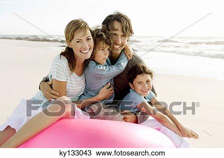 Stock Image - Parents and two children sitting on the beach, posing for the camera, outdoors. Fotosearch