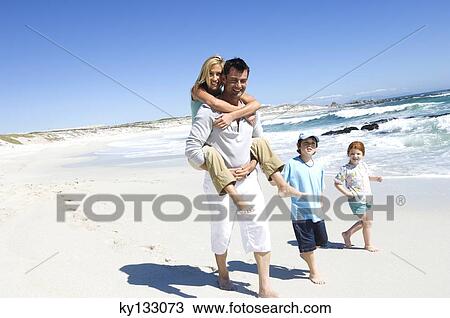 Stock Image - Parents and two children walking on the beach, man carrying woman on his back, outdoors. Fotosearch