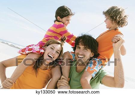 Stock Image - Portrait of parents carrying their two children on their shoulders, beach, outdoors. Fotosearch