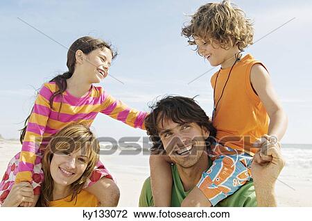 Stock Image - Portrait of parents carrying their two children on their shoulders, beach, outdoors. Fotosearch