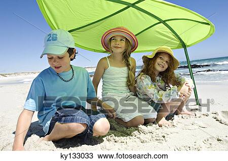 Stock Image - Three brother and sisters sitting on the beach, smiling, under a great green leaf, outdoors. Fotosearch
