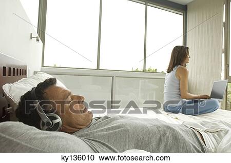 Couple in bedroom, man listening to music, woman using laptop, indoors View Large Photo Image Stock Image - Couple in bedroom, man listening to music, woman using laptop, indoors. Fotosearch