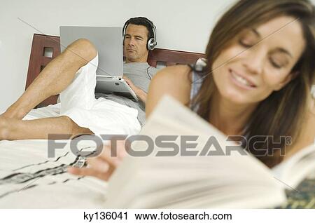 Couple on bed, woman reading, man using laptop, indoors View Large Photo Image Stock Image - Couple on bed, woman reading, man using laptop, indoors. Fotosearch