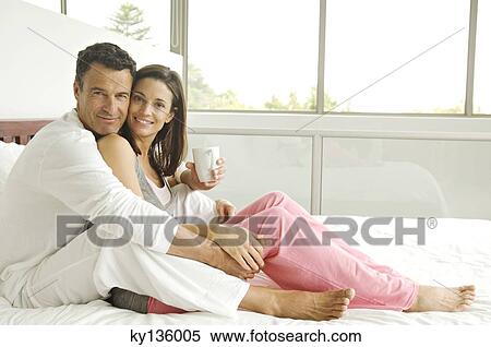 Couple sitting on bed, looking at the camera, indoors View Large Photo Image Stock Photography - Couple sitting on bed, looking at the camera, indoors. Fotosearch