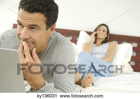 Man using laptop, woman phoning lying on bed in background, indoors View Large Photo Image Stock Image - Man using laptop, woman phoning lying on bed in background, indoors. Fotosearch
