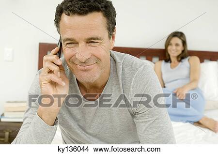 Portrait of a man phoning with cell, woman sitting on bed in background, indoors View Large Photo Image Picture - Portrait of a man phoning with cell, woman sitting on bed in background, indoors. Fotosearch