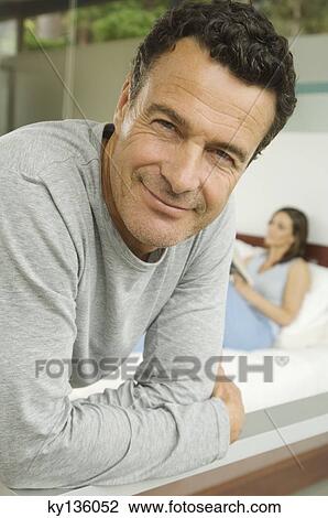 Portrait of a man posing for the camera, woman on bed in background, indoors View Large Photo Image Stock Image - Portrait of a man posing for the camera, woman on bed in background, indoors. Fotosearch