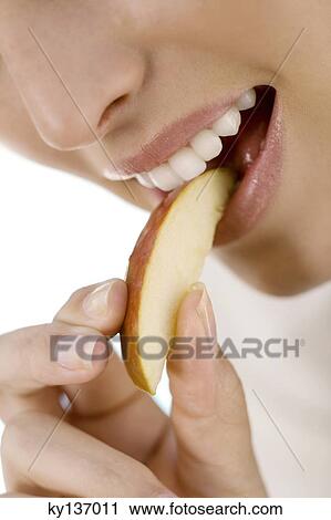 Stock Image - Portrait of a young woman eating a piece of apple, indoors. Fotosearch