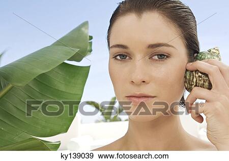 Portrait of a young woman holding a shell against her ear, outdoors View Large Photo Image Stock Photo - Portrait of a young woman holding a shell against her ear, outdoors. Fotosearch